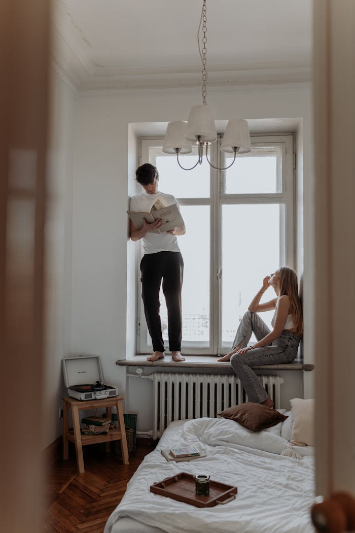 Two young adults enjoy a relaxed moment by the window, one reading while the other sits on a windowsill.